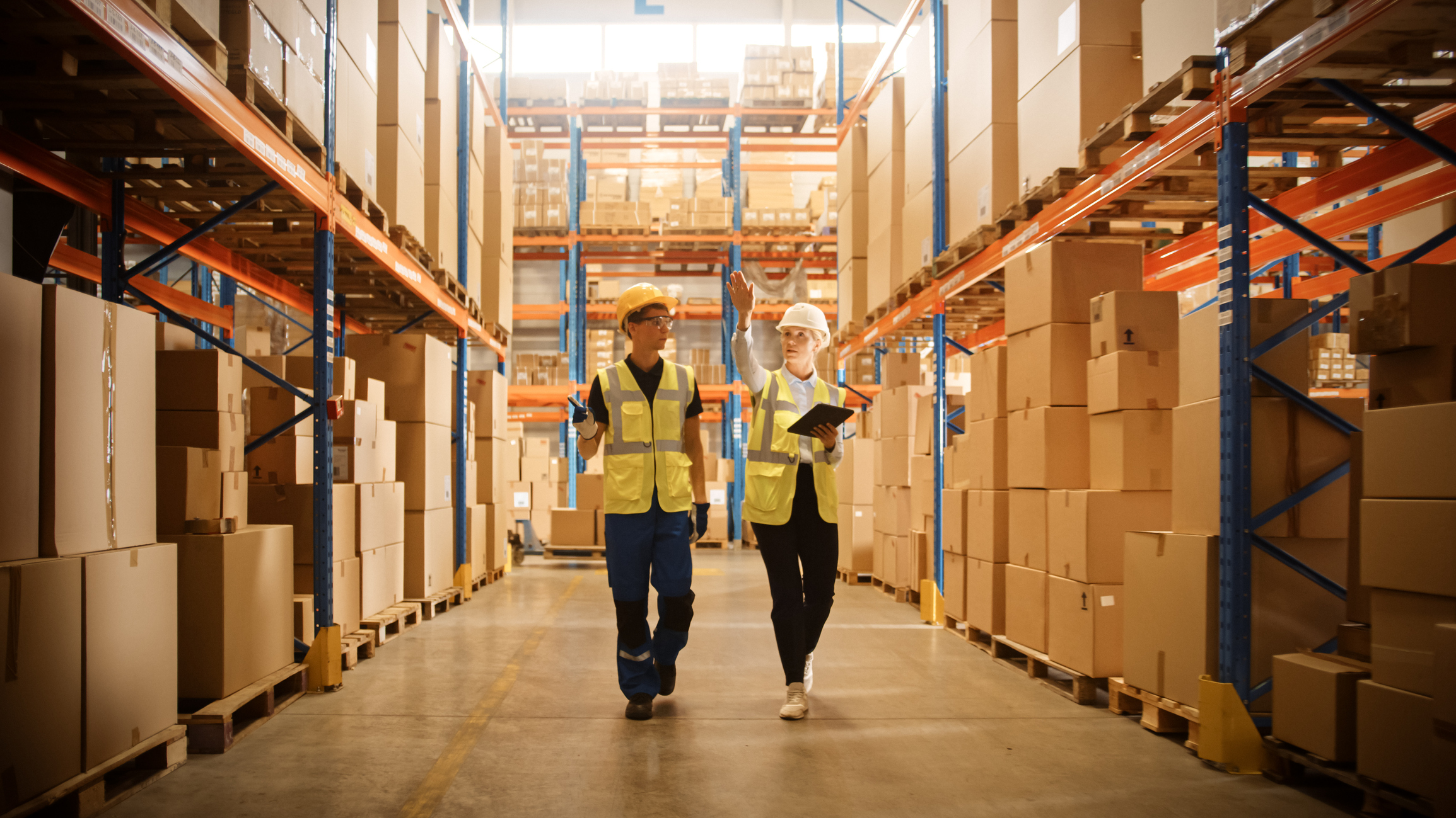 Retail Warehouse full of Shelves with Goods in Cardboard Boxes, Male Worker and Female Supervisor Holding Digital Tablet Discuss Product Delivery while Scanning Packages. Distribution Logistics Center